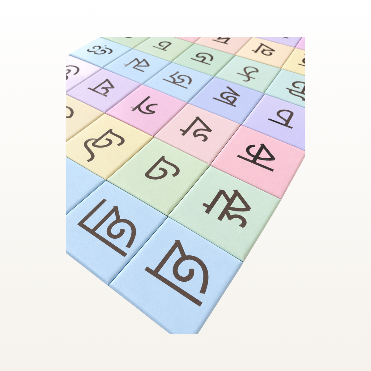 A set of pastel-colored square tiles arranged in a grid, each printed with a different Bangla script letter in dark brown. The tiles feature soft pastel shades of blue, green, pink, yellow, and lavender on a clean, off-white background.